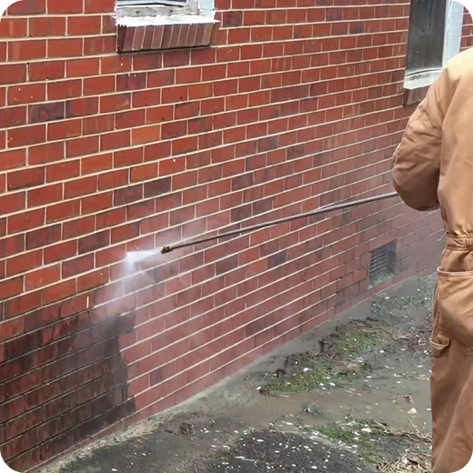 A worker pressure washing a red brick wall, removing dirt and grime.
