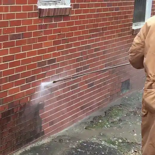 A worker pressure washing a red brick wall, removing dirt and grime.